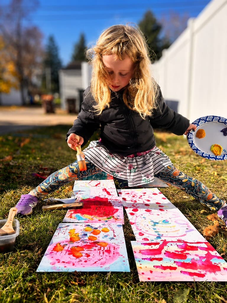A girl painting outdoors in autumn, crouching over several colorful abstract works spread on the grass