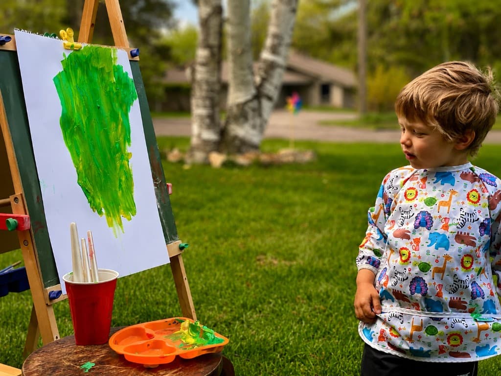 A young boy in an animal-print smock painting at an outdoor easel on a sunny lawn, quiet and focused on his green canvas
