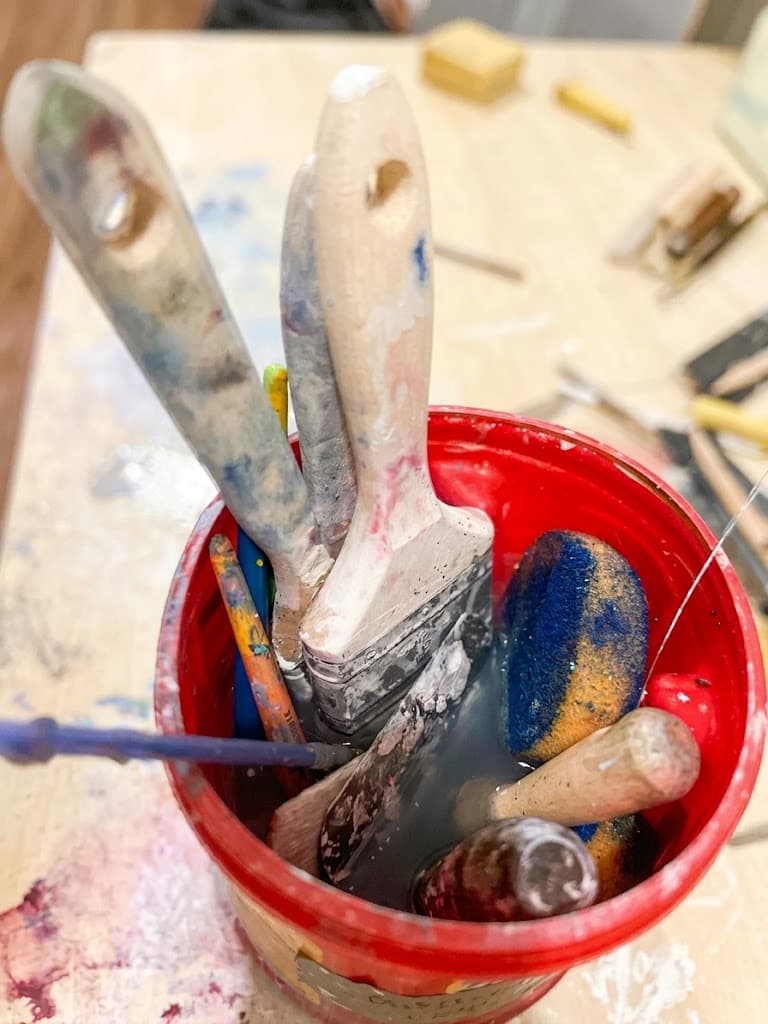 Overhead view into a well-used red brush bucket with paintbrushes, tools, and a sponge in cloudy rinse water