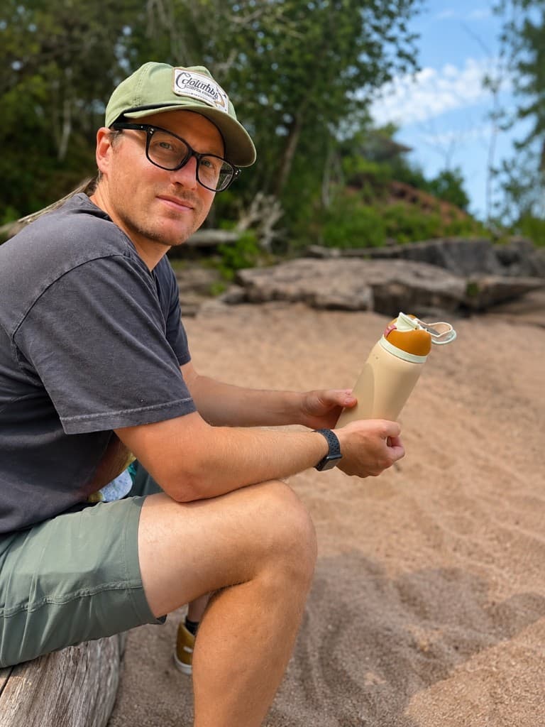 Michael Johnson sitting on a driftwood log at the beach, wearing a Columbia cap and holding a water bottle