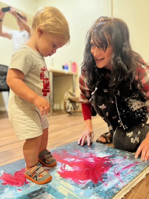 Rose kneeling at a toddler's eye level over a canvas on the floor, hands in blue paint, both smiling
