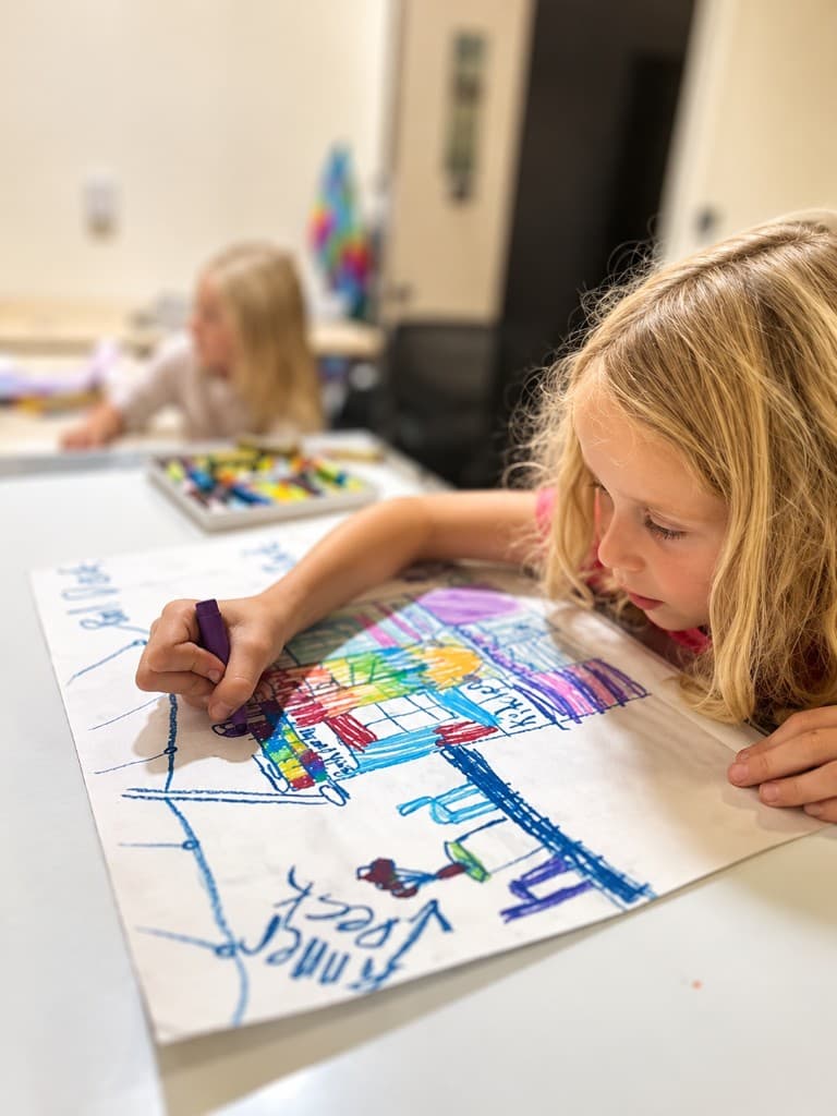 A young girl leaning close over her drawing, absorbed in colorful work at a sunlit studio table