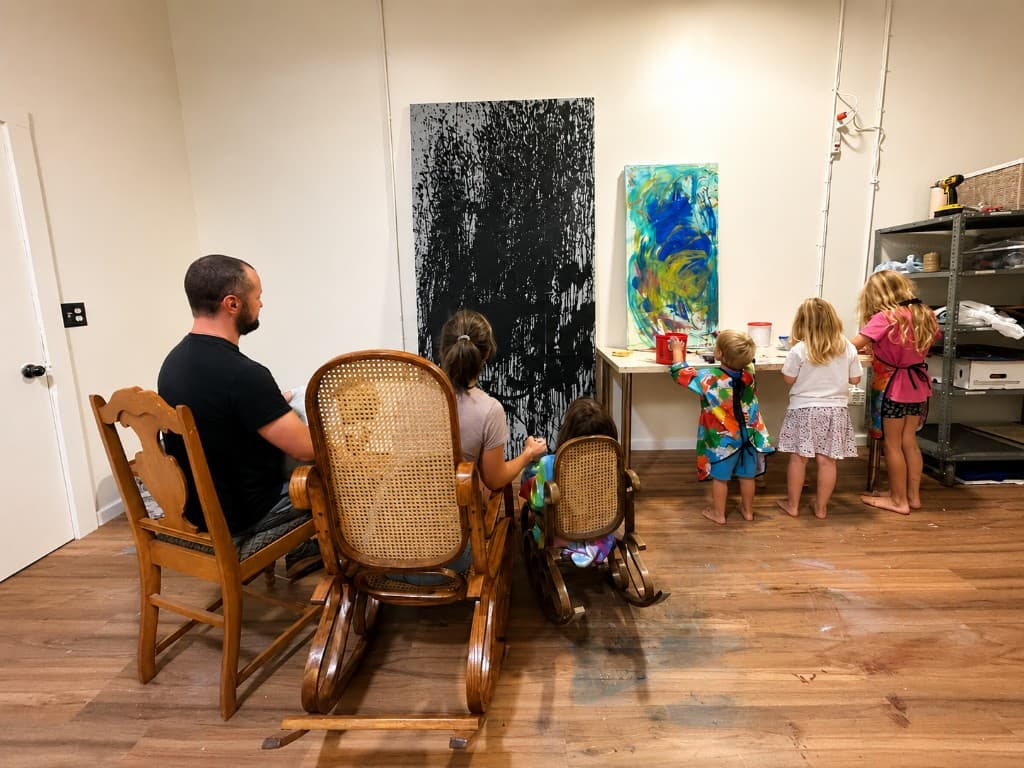 A family gathered in the studio — an adult and children seated in rocking chairs, other children working at a table with paint and materials, large abstract paintings on the wall behind them.
