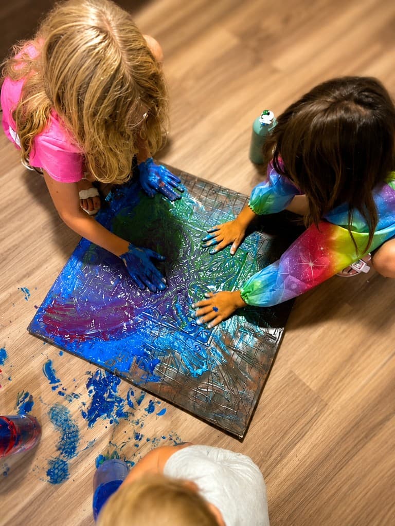 Three children with hands covered in blue and teal paint, working together on a collaborative floor painting