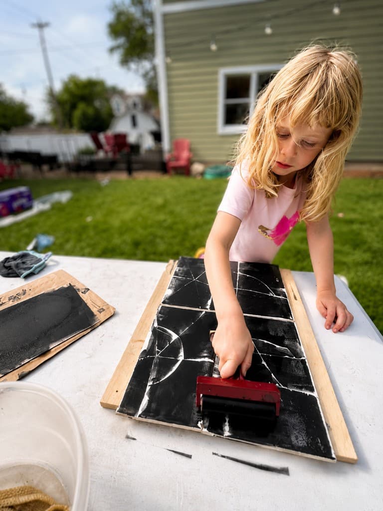 A girl in a pink shirt rolling ink on a monoprint board, with string lights and evening sky behind her