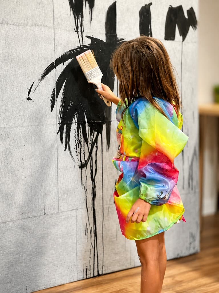 A girl in a rainbow smock making a large black gestural brushstroke on a wall-mounted panel, seen from behind