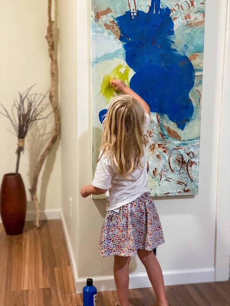 A young girl seen from behind, reaching up to paint on a large blue and teal canvas hanging on the studio wall